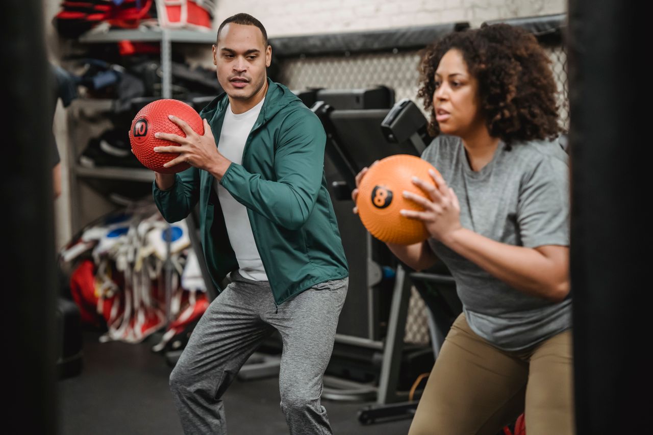 Serious young ethnic male instructor in sportswear looking at plus size African American female doing squat exercise with medicine ball in gym